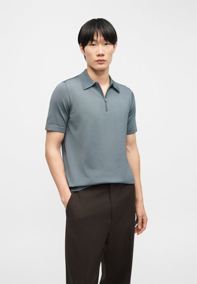 Νεαρός άνδρας με μαύρα μαλλιά στέκεται με τα χέρια στις τσέπες, φοράει μπλε κοντομάνικο πόλο με φερμουάρ και σκούρο καφέ παντελόνι.