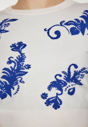 Maglione bianco lavorato a maglia con motivi floreali blu e a spirale. Vista ravvicinata della texture e dei dettagli del design sulla parte anteriore.