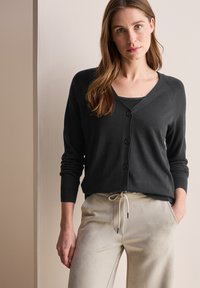 Cardigan noir à boutonnage avec un col en V, des manches longues et des poignets côtelés. Porté par-dessus un débardeur noir, associé à un pantalon à cordon de serrage beige.
