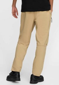 Beżowe spodnie wykonane z lekkiego materiału, o prostym kroju, z suwakiem w kieszeni i elastycznymi mankietami, zestawione z czarnymi trampkami.