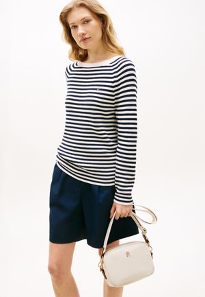 Maglione a righe lavorato a maglia in blu navy e bianco, abbinato a pantaloncini blu navy. Il modello tiene in mano una piccola borsa crema con dettagli metallici dorati.