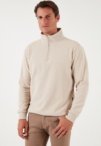 Bež flis pulover s visokim ovratnikom, polu-zip prednjim dijelom, rebrastim manžetama i glatkom teksturom. Minimalistički dizajn bez vidljivih uzoraka ili logoa.