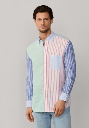 Homme portant une chemise à manches longues boutonnée avec des rayures verticales vertes, rouges et bleues, associée à un jean bleu clair.