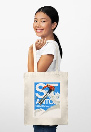 Lächelnde Frau mit schwarzen Haaren hält eine beige Tragetasche, auf der ein Skifahrer mit Stöcken vor blauem Himmel und Schnee springt, mit dem Text „St. Anton am Arlberg.“