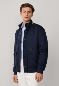 Chaqueta acolchada de color azul marino con cuello alzado, cierre de botón, bolsillos delanteros y puños acanalados. Tejido ligero con textura suave.