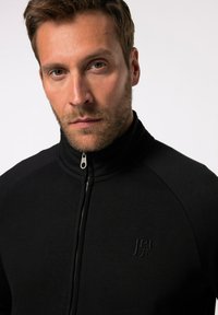 Sort zip-up jakke lavet af blødt materiale med høj krave, med et præget logo på brystet og en sølv lynlåsdetalje.