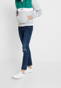 Sudadera con cremallera gris y verde, con un bolsillo tipo canguro, acompañada de un par de jeans oscillos azules con detalles desgastados, y zapatillas blancas de fácil puesta.
