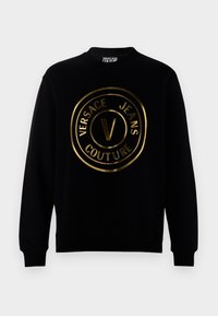 Črna dukserica z okroglo zlatim logotipom, ki vsebuje besedilo "VERSACE JEANS COUTURE" in izrazit stiliziran "V" na sredini.