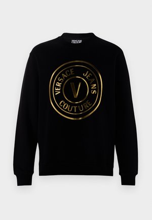 Melnā sporta veste ar apaļu zeltītu logo, kurā ir teksts "VERSACE JEANS COUTURE" un izteikts stilizēts "V" centrā.