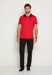Rotes Poloshirt mit schwarzem Kragen und Manschetten, kurzen Ärmeln und drei Knöpfen. Kombiniert mit schwarzen Hosen und schwarzen Schuhen mit roten Akzenten.