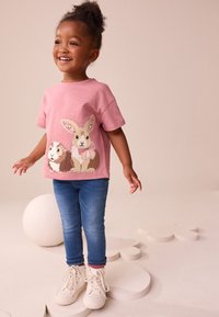 T-shirt rose à manches courtes avec un design de lapin et de hérisson, associé à un jean bleu et des baskets montantes blanches. Coupe avec un col rond.