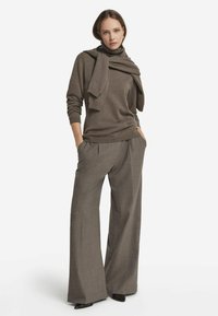 Kvinna som bär en brun polotröja med en matchande tröja över axlarna, vida grå byxor och svarta spetsiga skor.