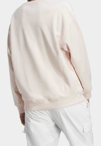 Světle růžová oversized mikina s žebrovanými manžetami a lemem, zdobená třemi bílými pruhy podél švu na ramenou. Materiál směs bavlny.