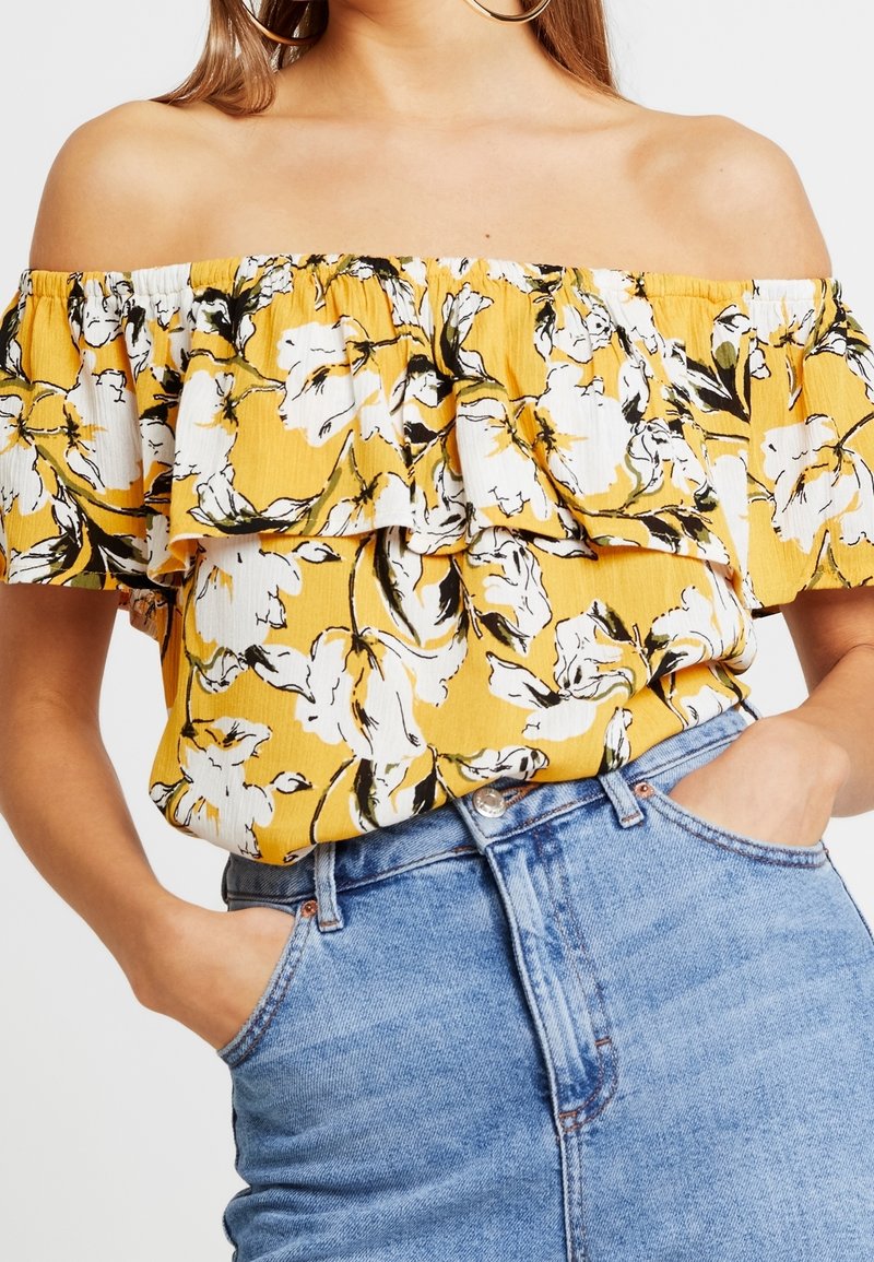 Haut jaune décolleté épaule avec imprimé floral blanc et noir, rentré dans un jean en denim bleu taille haute, mains dans les poches.