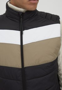 Schwarwest, beige und weiße gepolsterte Weste mit hohem Kragen und Reißverschluss. Auffälliges Zickzackmuster über der Brust. Gestepptes Textil auf den Schultern.
