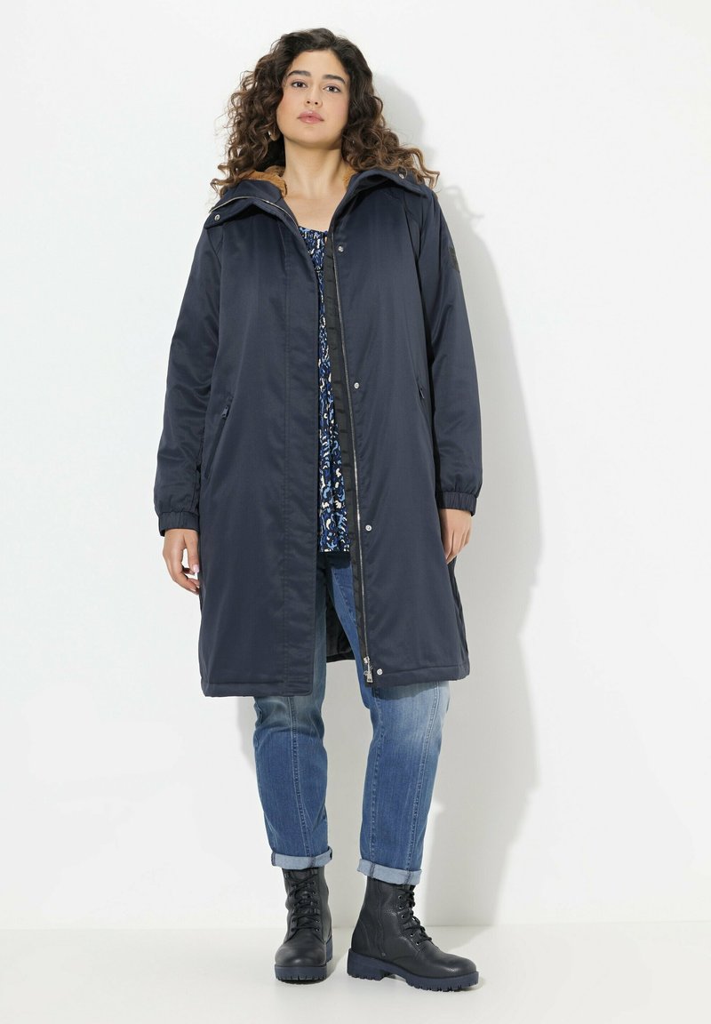 Marinblå lång parka med huva, med dragkedja fram, sidofickor och elastiska ärmslut. Buren över en mönstrad topp och jeans, med snörade stövlar.