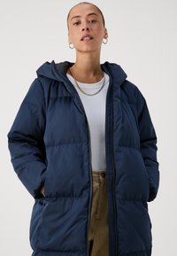 Tumši zilas krāsas pūkains jaka ar kapuci, ar rāvējslēdzēja aizdari, divām sānu kabatām un quiltēta dizaina. Valkāta pār gaiši pelēku kreklu.