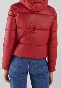 Rød puffjakke med hætte, der har et quiltet design og synlige sømme. Skåret lidt over taljen, parret med blå jeans.