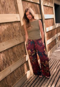 Débardeur olive tricoté et pantalon bordeaux ample à motif avec détails floraux. Debout contre un mur en bois. Du sable sous les pieds.