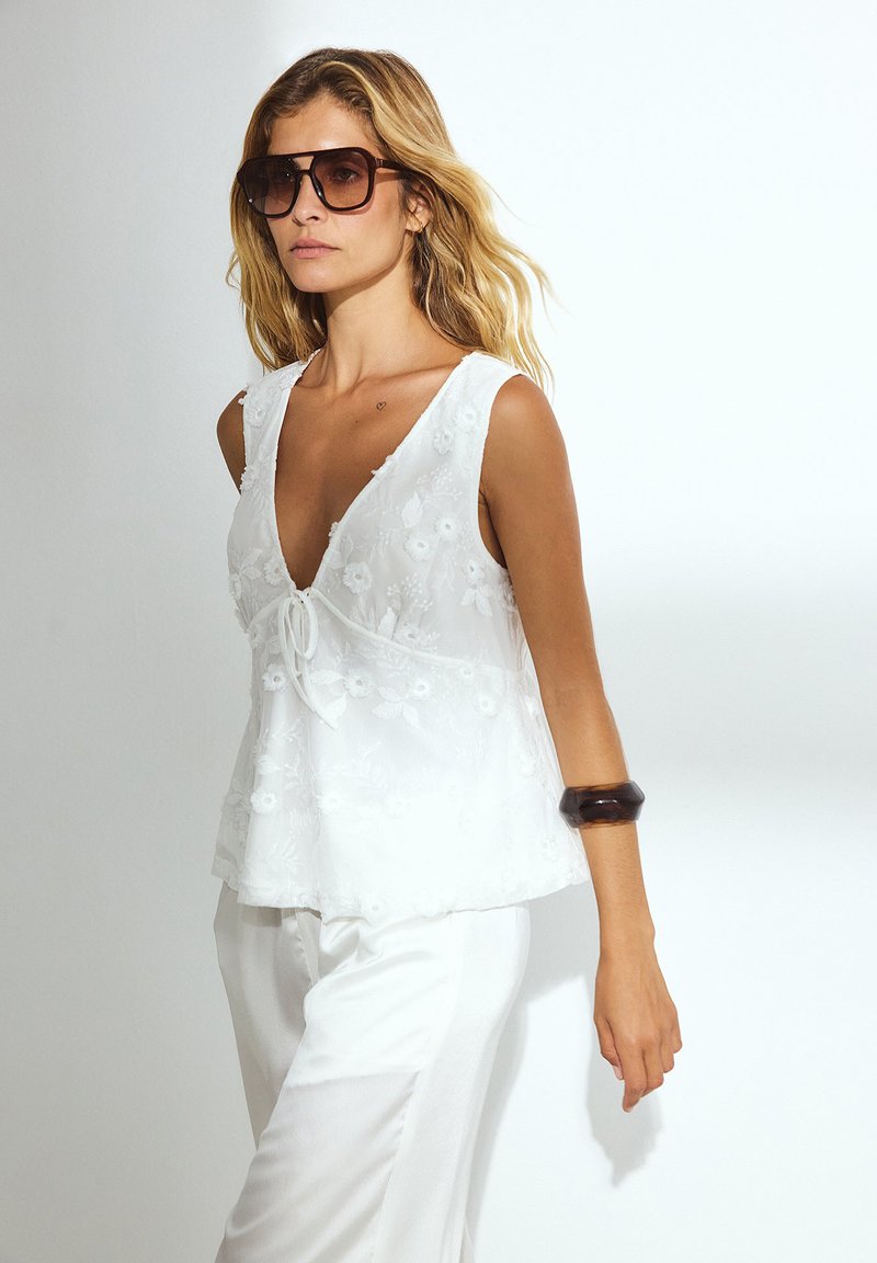 Witte mouwloze top met bloemembroiderie, V-hals en een strikdetail in de taille. Draagt bij witte broeken en een stevige bruine armband.