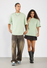 Světle zelené oversized trička, zkombinovaná s vybledlými hnědými širokými džíny a černou džínsovou sukní. Modelka má na sobě černé kotníkové boty se sponami.