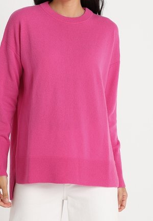 Rosa gestrickter Pullover mit rundem Halsausschnitt, überschnittenen Schultern und geripptem Saum. Weicher Stoff und lässige Passform, kombiniert mit weißen Hosen.
