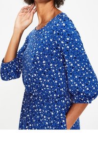 Blåblommig klänning med rund halsringning, trekvartsärmar och insvängd midja. Tyget pryds av vita blommor på en blå bakgrund.