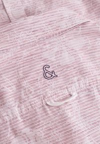 Tessuto rosa chiaro con un sottile motivo floreale e sottili righe rosse. Presenta un logo ricamato di colore blu scuro con la &. Include un piccolo dettaglio a cerchio.