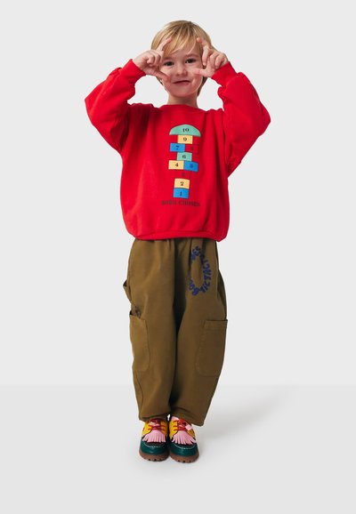 Felpa rossa con un design di campana giocoso, pantaloni baggy verde oliva con tasche laterali e scarpe colorate con dettagli in frange.