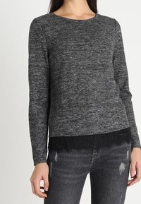 Svetr s dlouhým rukávem a kulatým výstřihem v melírovaně šedé barvě, s černou krajkovou lemem. Nošený s roztrhanými černými džínami, které zdůrazňují texturu látky.