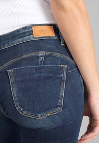 Osoba v tmavě modrých denimových džínách s viditelnou zadní kapsou a koženou značkovou etiketou, stojící na neutrálním pozadí.