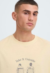 T-shirt en coton couleur crème avec une impression graphique de deux silhouettes assises sur des chaises, accompagnée du texte « Take It Outside ». Coupe classique à col rond.