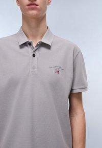 Šedé polo tričko vyrobené z texturované tkaniny. Obsahuje límeček, tři knoflíky a malé vyšívané logo s vlajkou na prsou.
