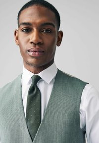 Green-textured vest over a white shirt, paired with a dark green tie featuring small white dots. The background is neutral.