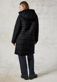 Svart quiltad pufferjacka med huva, som har en lång silhuett, diagonala sömmar och dragkedjor vid ärmslut. Bärs över mörka jeans och stövlar.