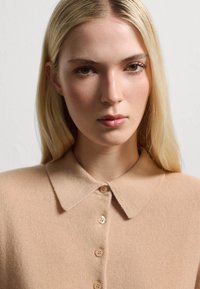 Beige gebreide trui met een kraag en een sluiting met vijf knopen. Zachte textuur, lichtgewicht en subtiele patroon details in het breiwerk.