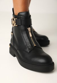 Botas de tobillo de cuero negro con una cremallera dorada, correa decorativa que presenta un logo dorado y una suela gruesa de goma negra.