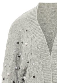 Svetle šedý pletený cardigan s kabelovým vzorem, doplněný černobílými perlami na rukávech. Design s otevřeným předním dílem.