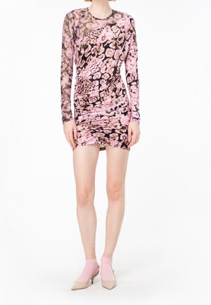 Kobieta w różowej i czarnej mini sukience z marszczeniami oraz przezroczystymi długimi rękawami, w różowych skarpetkach i beżowych butach na niskim obcasie.