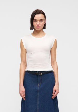 Jeune femme aux cheveux courts et foncés, portant un haut blanc sans manches, des boucles d'oreilles créoles, une ceinture noire et une jupe en jean bleue, debout face à l'avant.