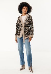Beige en bruine cardigan met luipaardprint, vastgebonden in de taille, gecombineerd met lichtblauwe denim jeans met een split aan de onderkant, en metallic hakken.