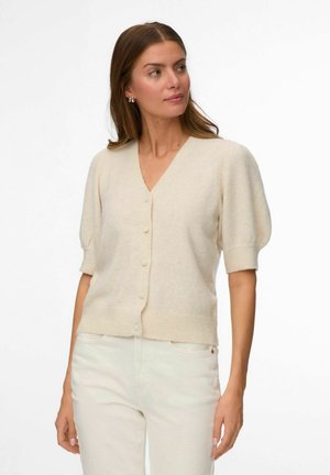 Vrouw met lichtbruin haar, gekleed in een crèmekleurige korte mouw gebreide cardigan met knopen en witte broek, kijkt naar links tegen een effen achtergrond.