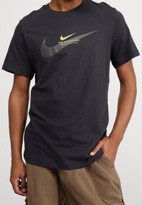 Černé bavlněné tričko s velkým logem Nike ve tmavě šedé a zlaté barvě, s textem "Nike Athletic Dept." ve žluté barvě pod ním.