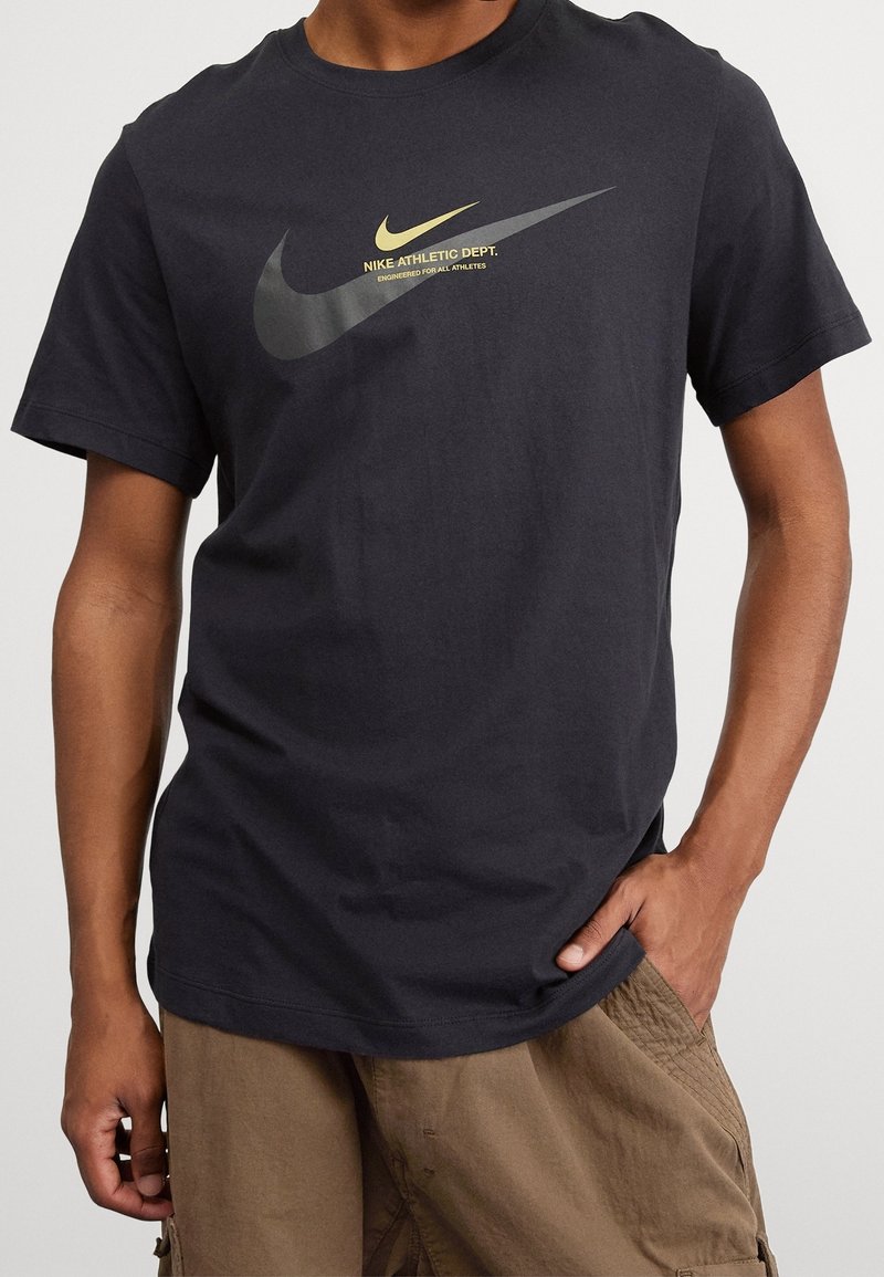 Černé bavlněné tričko s velkým logem Nike ve tmavě šedé a zlaté barvě, s textem "Nike Athletic Dept." ve žluté barvě pod ním.