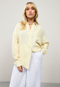 Yellow button-up shirt made of textured fabric with a patch pocket, paired with white linen pants, featuring long sleeves and a relaxed fit.