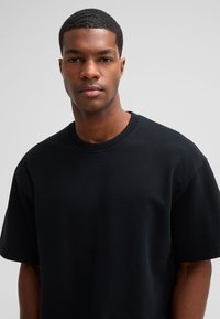 Zwarte korte mouwen crew neck t-shirt met een gestructureerde afwerking, voorzien van een ontspannen pasvorm en stevige naden. Achtergrond is neutraal grijs.