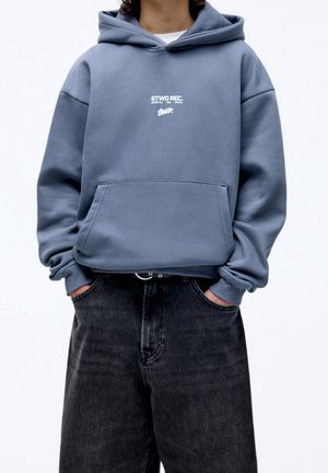Personne portant un sweat à capuche bleu avec le texte "STWD REC." et un jean noir, les mains dans les poches, debout devant un fond blanc uni.