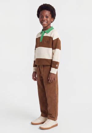 Dreng står og smiler iført en brun og cremefarvet stribet sweater med grøn krave, brune kordbuksbukser og hvide sneakers.