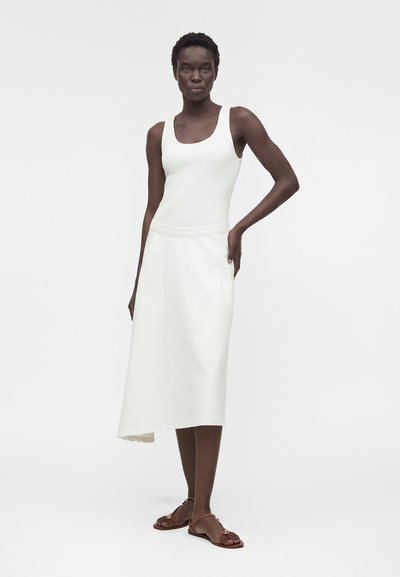 Γυναίκα με λευκό, αμάνικο τοπ με ριπ ύφανση, λευκή ασύμμετρη μίντι φούστα και καφέ επίπεδα σανδάλια, στέκεται με το ένα χέρι στη μέση.