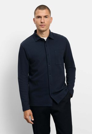 Uomo che indossa una camicia blu scuro a maniche lunghe con taschino sul petto, in piedi davanti a uno sfondo grigio chiaro.